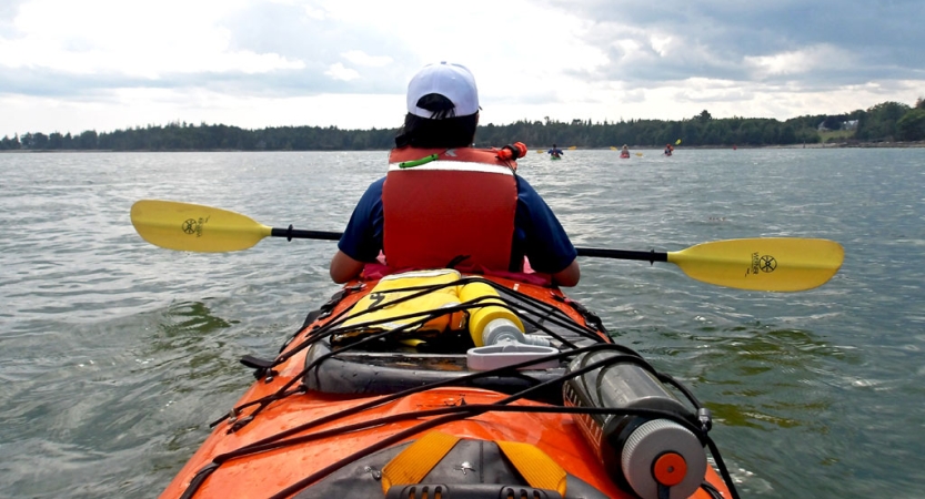 From the back of an orange kayak, a person wearing a life jacket holds a yellow paddle and looks out over the calm water. 